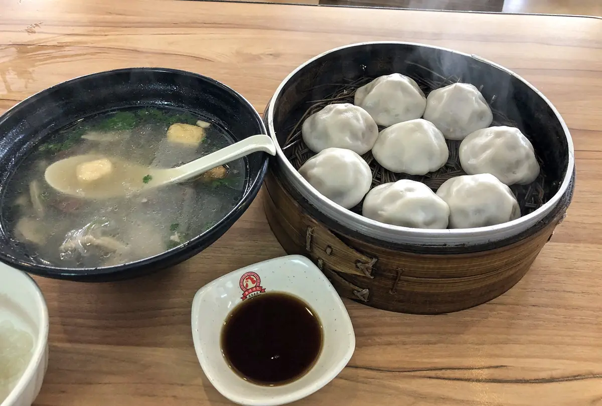 A steamer basket of soup dumplings next to a bowl of duck blood vermicelli soup